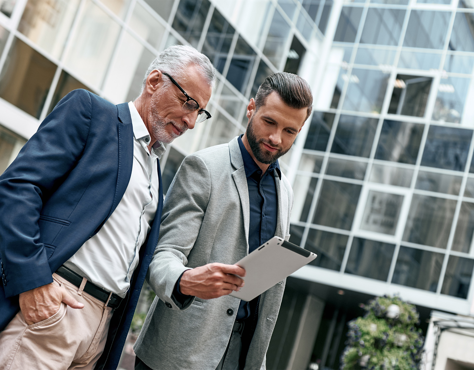 Zwei Business Professionals in modernen Anzügen betrachten ein Tablet vor einem gläsernen Bürogebäude – repräsentativ für die Beratungs- und Investmenttätigkeit von ENIGMA GmbH, die auf Immobilieninvestments, Venture Capital und Photovoltaik spezialisiert ist.